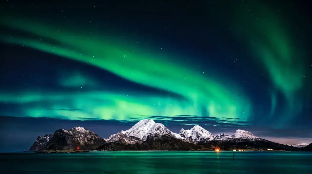 Tromsø nocą: zielona zorza polarna tańczy nad ośnieżonymi górami i spokojnym morzem.