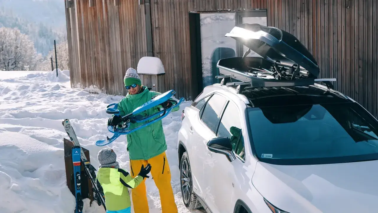 Rodzina przygotowuje się do wyjazdu na narty. Na dachu białego SUV-a zamontowany jest box dachowy, idealny na sprzęt zimowy.