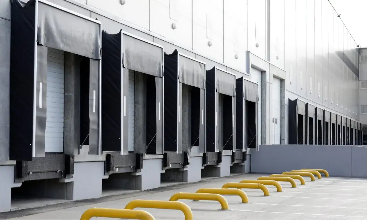 Rząd dok&oacute;w przeładunkowych z czarnymi fartuchami i białymi drzwiami rolowanymi. Przed nimi ż&oacute;łte odbojniki.