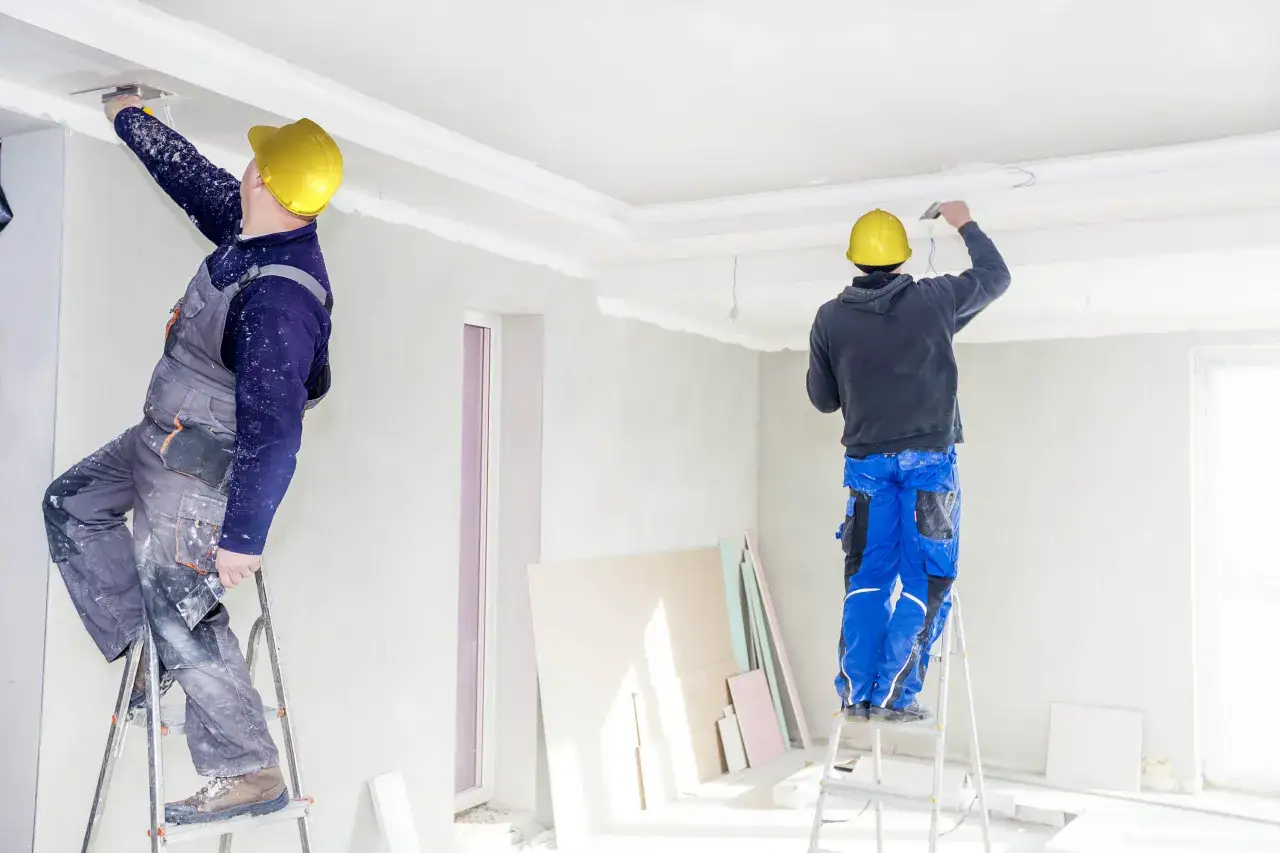 Dw&oacute;ch budowlańc&oacute;w w ż&oacute;łtych kaskach wykonuje nakładanie akrylu przy suficie, stojąc na drabinach.