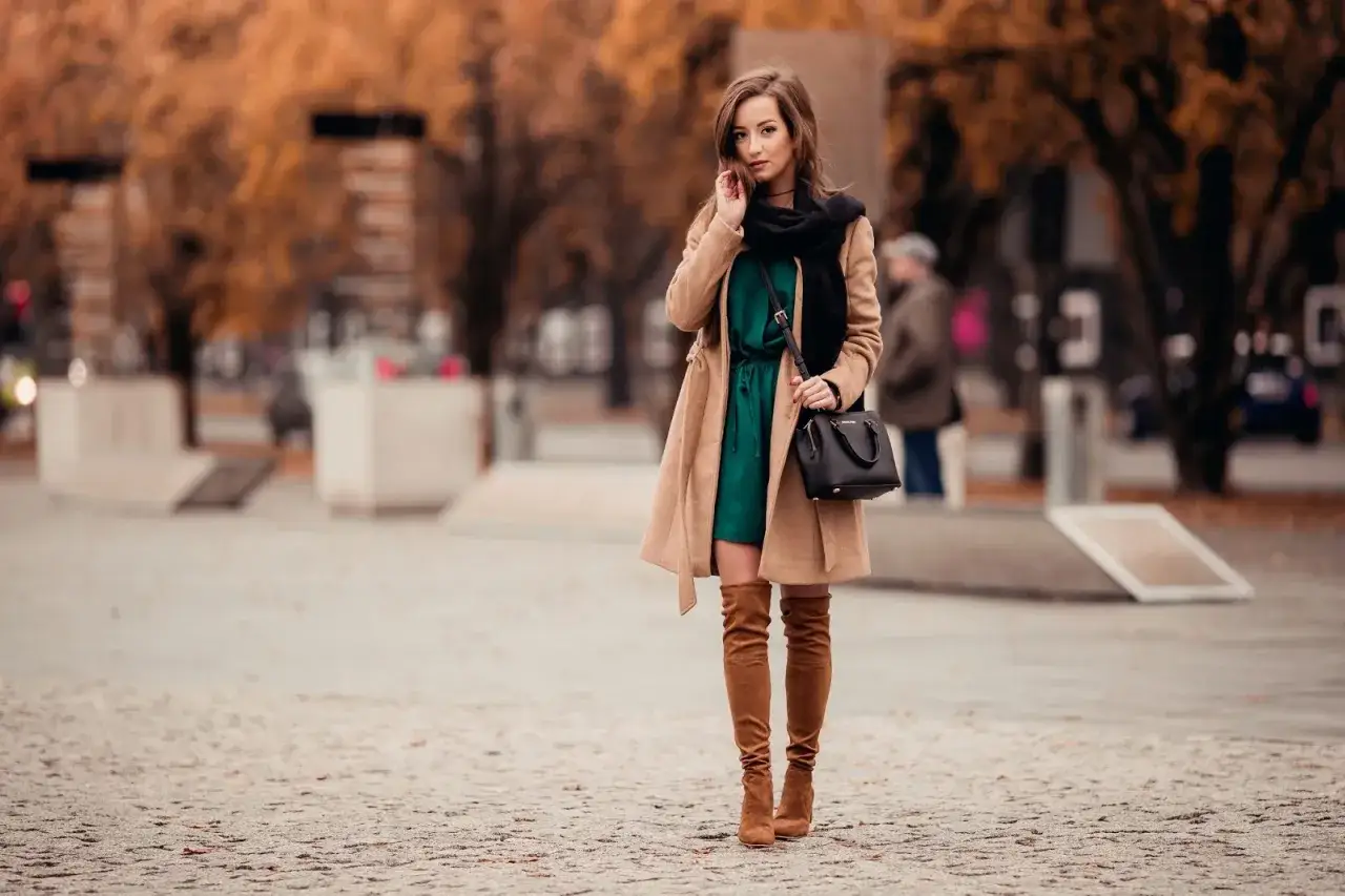 Elegancka kobieta w beżowym płaszczu i zielonej sukience, prezentująca stylowe karmelowe kozaki. Idealne karmelowe kozaki stylizacje na jesień.