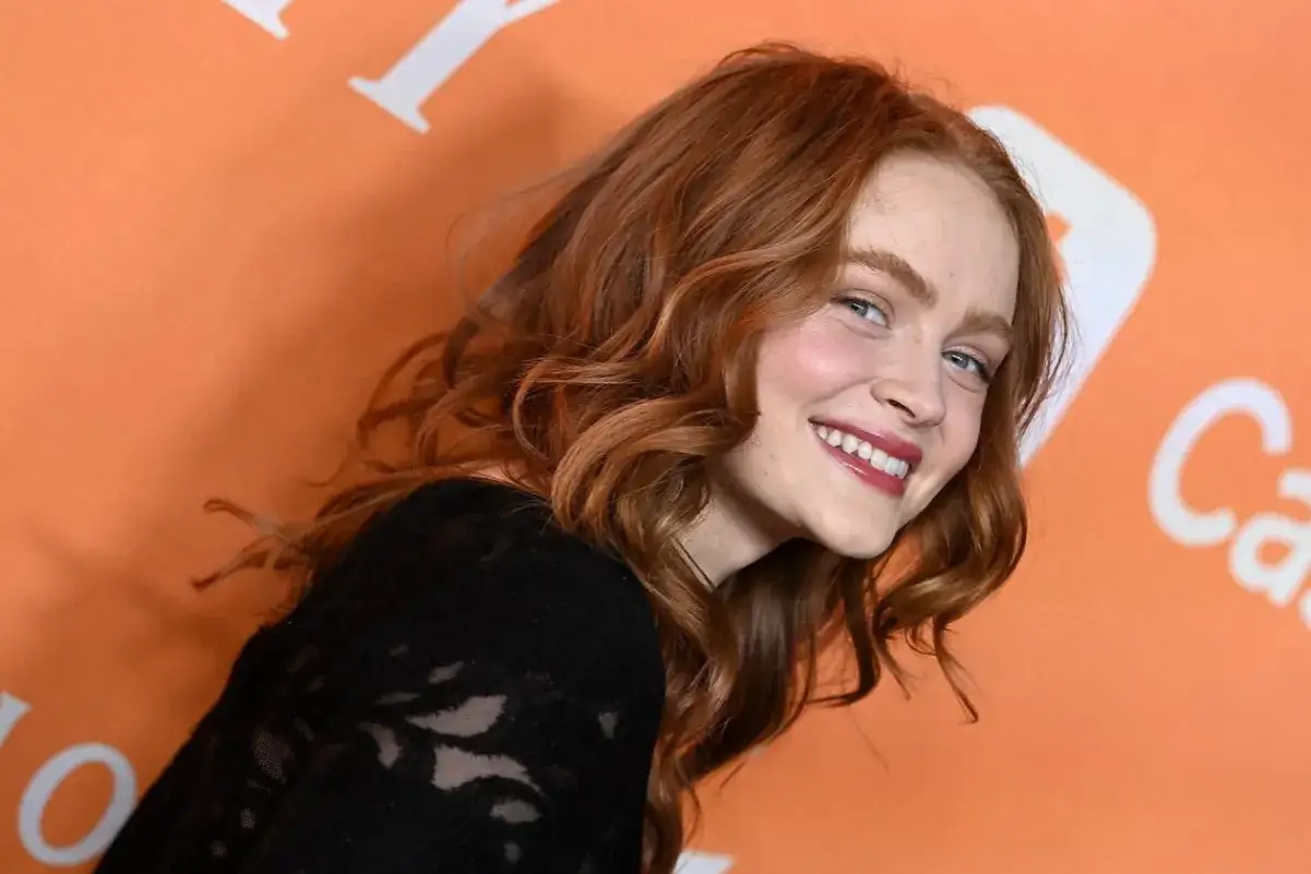 Uśmiechnięta Sadie Sink z rudymi włosami, znana z tego, w jakich filmach grał Sadie Sink, pozuje na tle pomarańczowego banera.