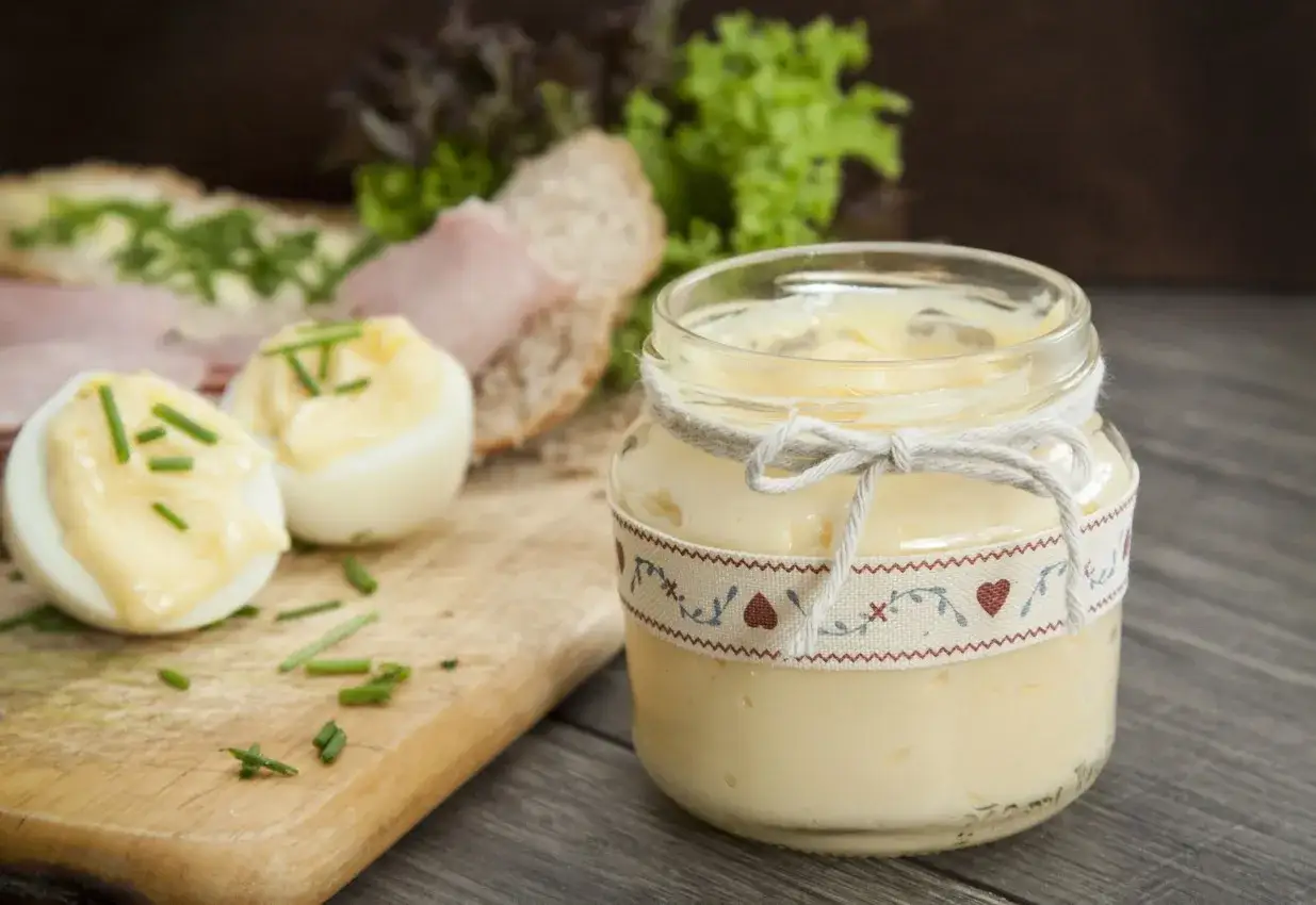 Słoik z domowym majonezem, obok jajka na twardo z majonezem i szczypiorkiem, na desce.