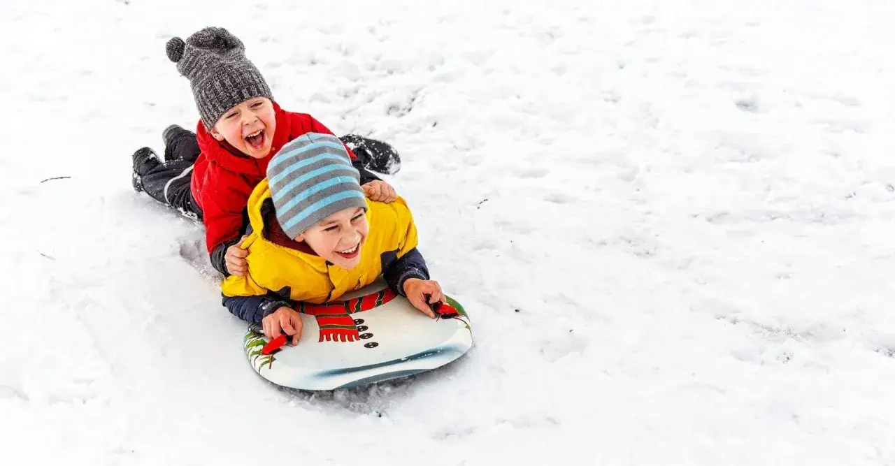 Dw&oacute;ch chłopc&oacute;w śmieje się radośnie, zjeżdżając na sankach po śniegu. Idealny dzień, by wybrać się na sanki!
