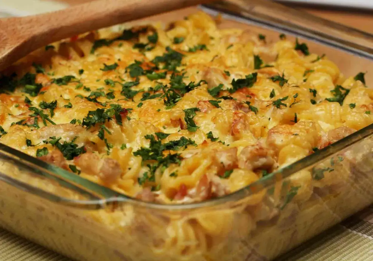 Pyszna zapiekanka makaronowa z kurczakiem, posypana serem i natką pietruszki, w szklanym naczyniu.