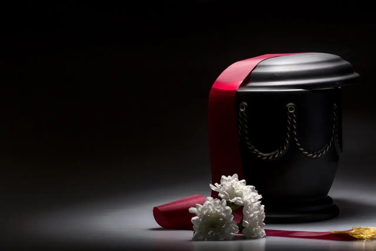 Czarna urna z czerwoną wstęgą i białymi kwiatami. Czy kremacja boli zmarłego? To pytanie, na które nauka daje jasną odpowiedź.