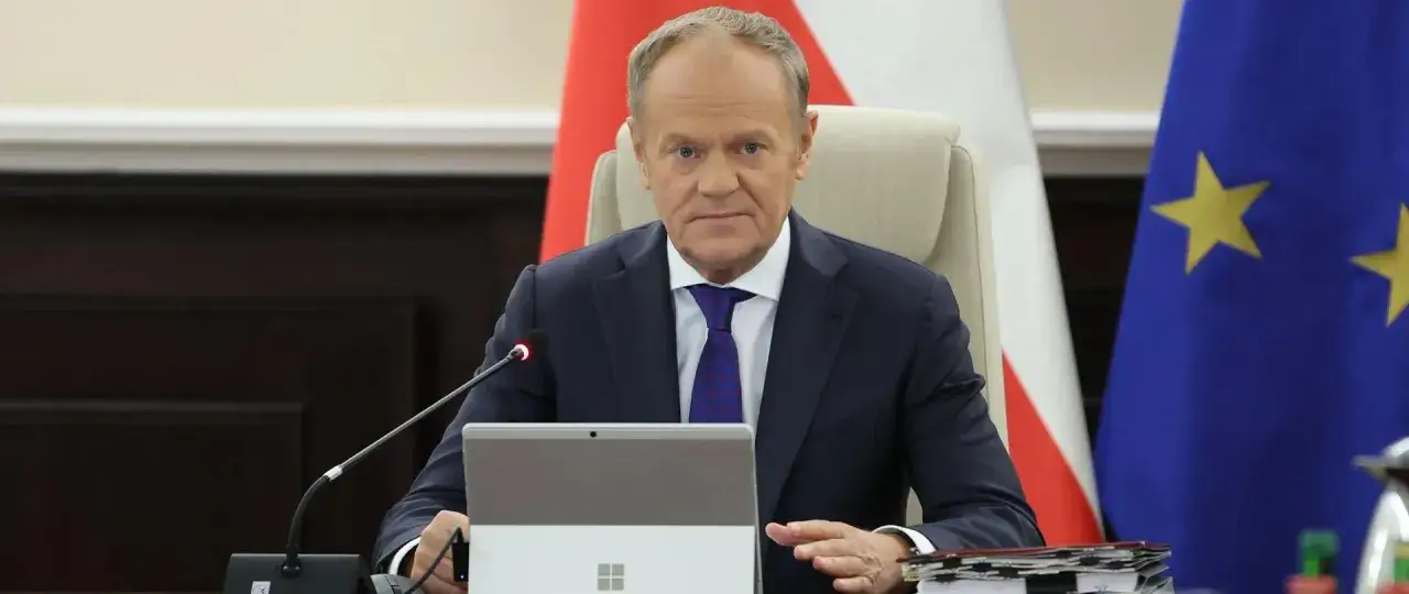 Donald Tusk dzisiaj podczas spotkania, siedzi przy stole z laptopem i stosem dokumentów. W tle flagi Polski i Unii Europejskiej.