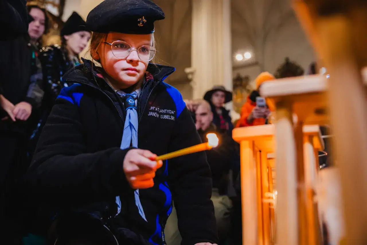 Dziewczynka w harcerskim mundurku i beretce zapala świecę. W tle widać innych uczestnik&oacute;w, być może podczas gry harcerskiej w pomieszczeniu.