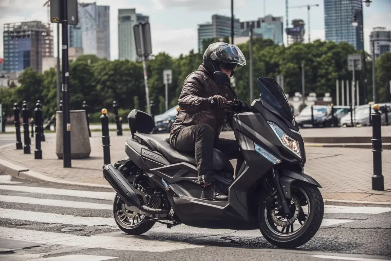 Mężczyzna w sk&oacute;rzanej kurtce jedzie na skuterze Zipp QR Max 125 przez miejskie przejście dla pieszych.