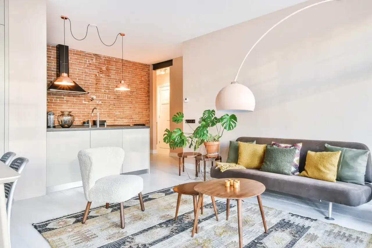 Stylowa aranżacja kawalerki 27m: kuchnia z ceglaną ścianą, przytulny salon z sofą i stolikami kawowymi, biały fotel.
