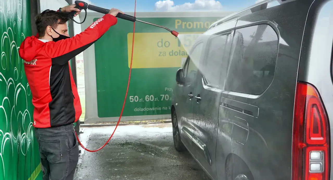 Mężczyzna w czerwonej bluzie pokazuje, jak myć auto na myjni, spryskując je wodą pod ciśnieniem.