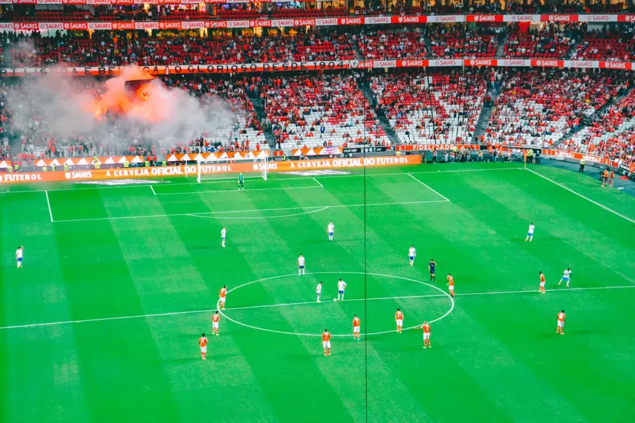 Stadion pełen kibiców klubu piłkarskiego z Lizbony. Na trybunach widać race, a na murawie toczy się mecz.