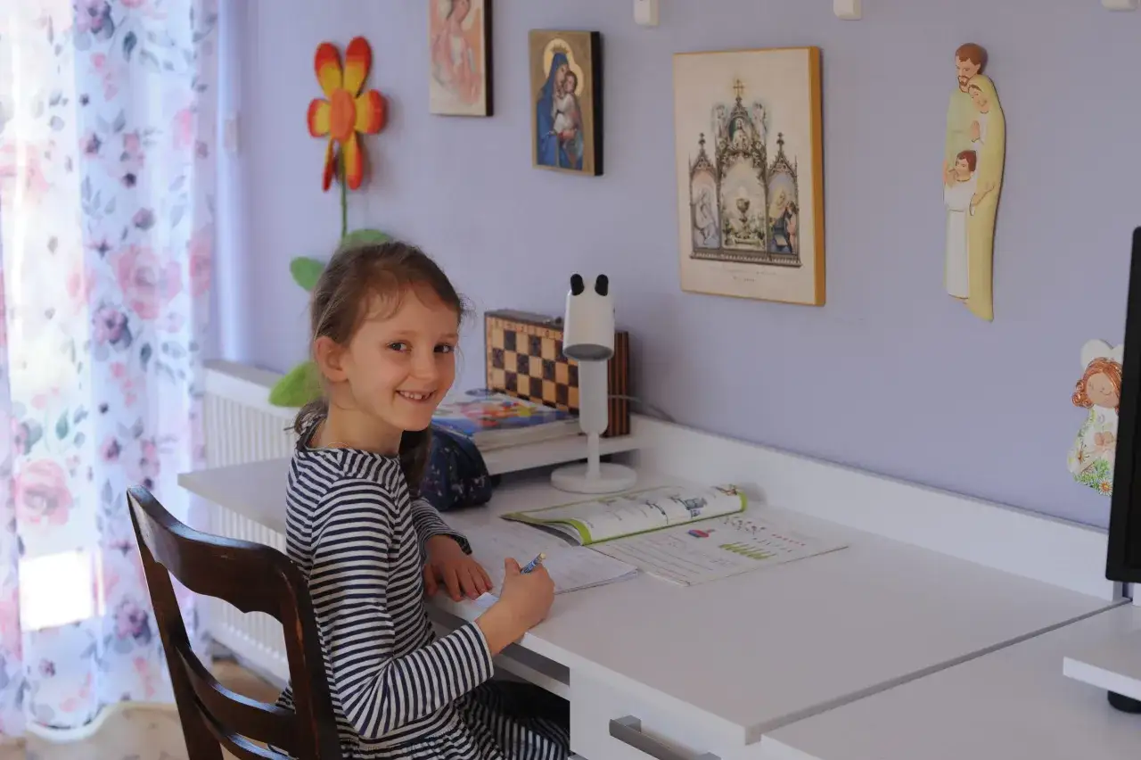 Uśmiechnięta dziewczynka w paski siedzi przy biurku, pisząc w zeszycie. To jej nauczanie domowe w podstawówce.