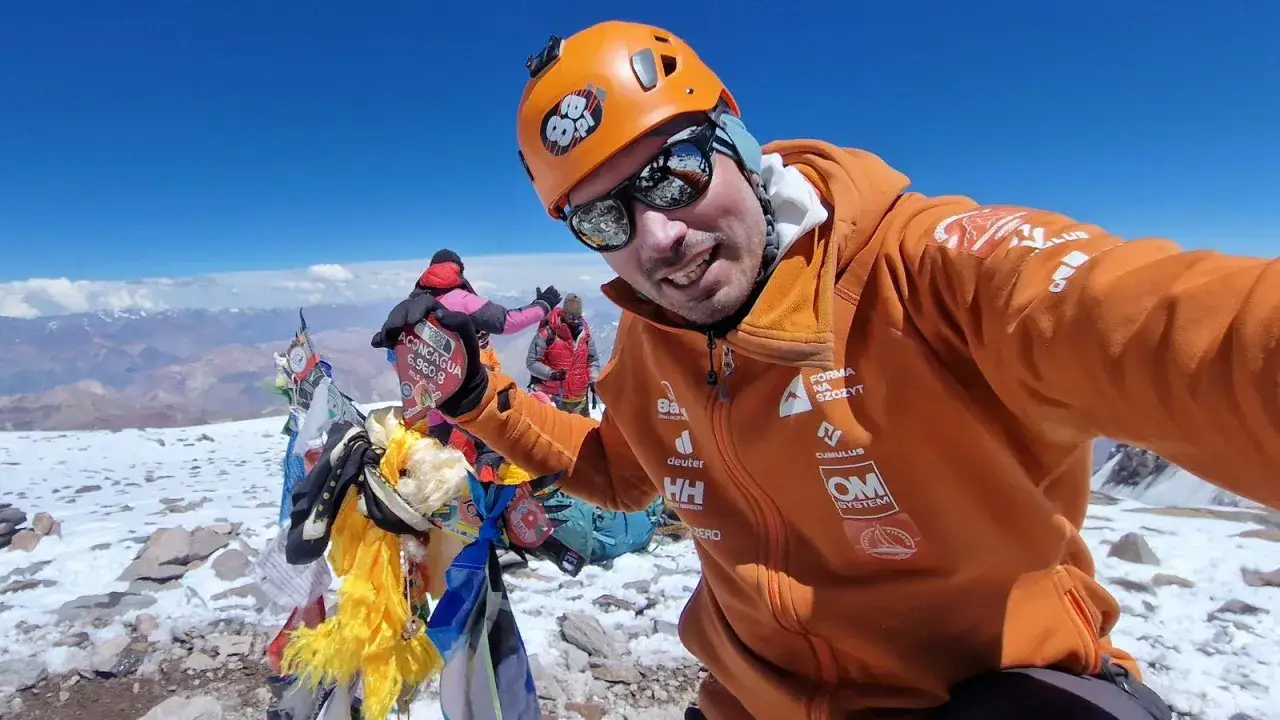 Szczęśliwy alpinista na szczycie Aconcagua, z flagami i innymi zdobywcami w tle.