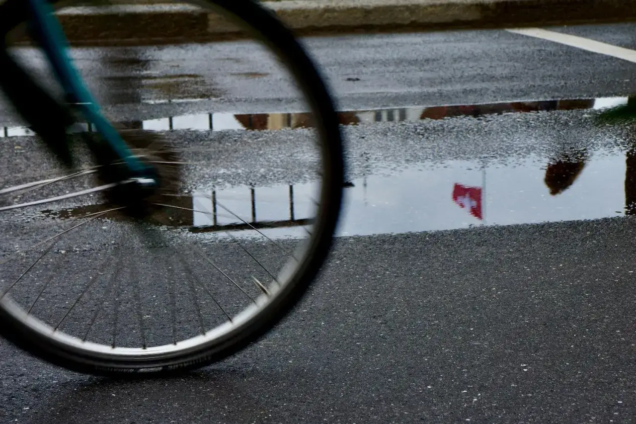 Koło roweru elektrycznego w deszczu odbija się w kałuży, na kt&oacute;rej widać szwajcarską flagę.
