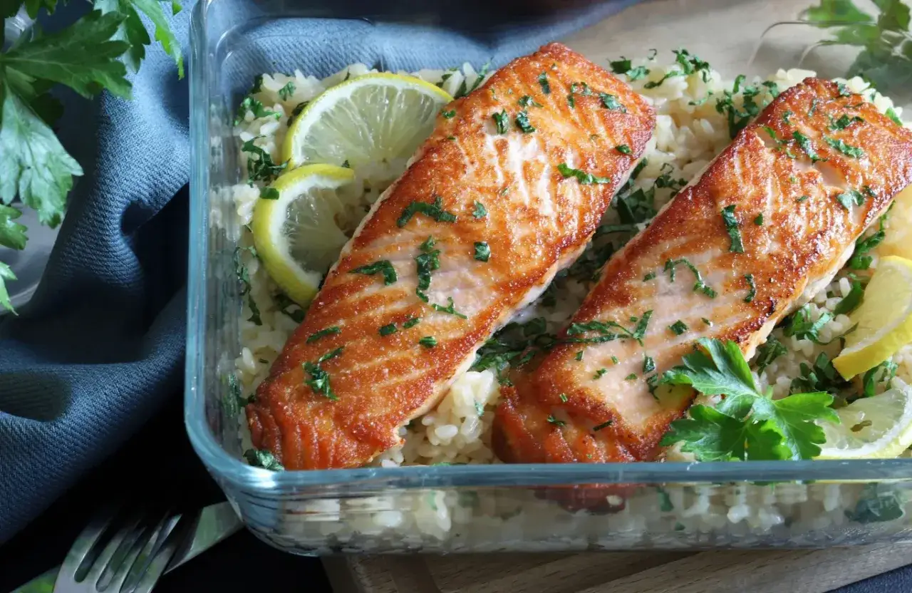 Dwa soczyste filety łososia na ryżu z cytryną i natką pietruszki w szklanym pojemniku. Pyszny posiłek gotowy do zabrania.