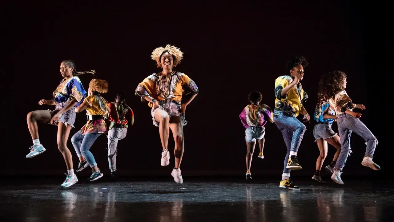 Grupa tancerzy w kolorowych strojach wykonuje energiczny układ choreograficzny. Scena jak z seriali o tańcu.