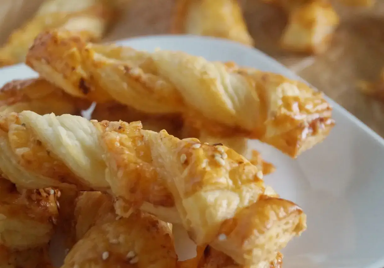 Złociste paluchy serowe z ciasta francuskiego, posypane sezamem, kuszą chrupkością i aromatem. Idealne na przekąskę.