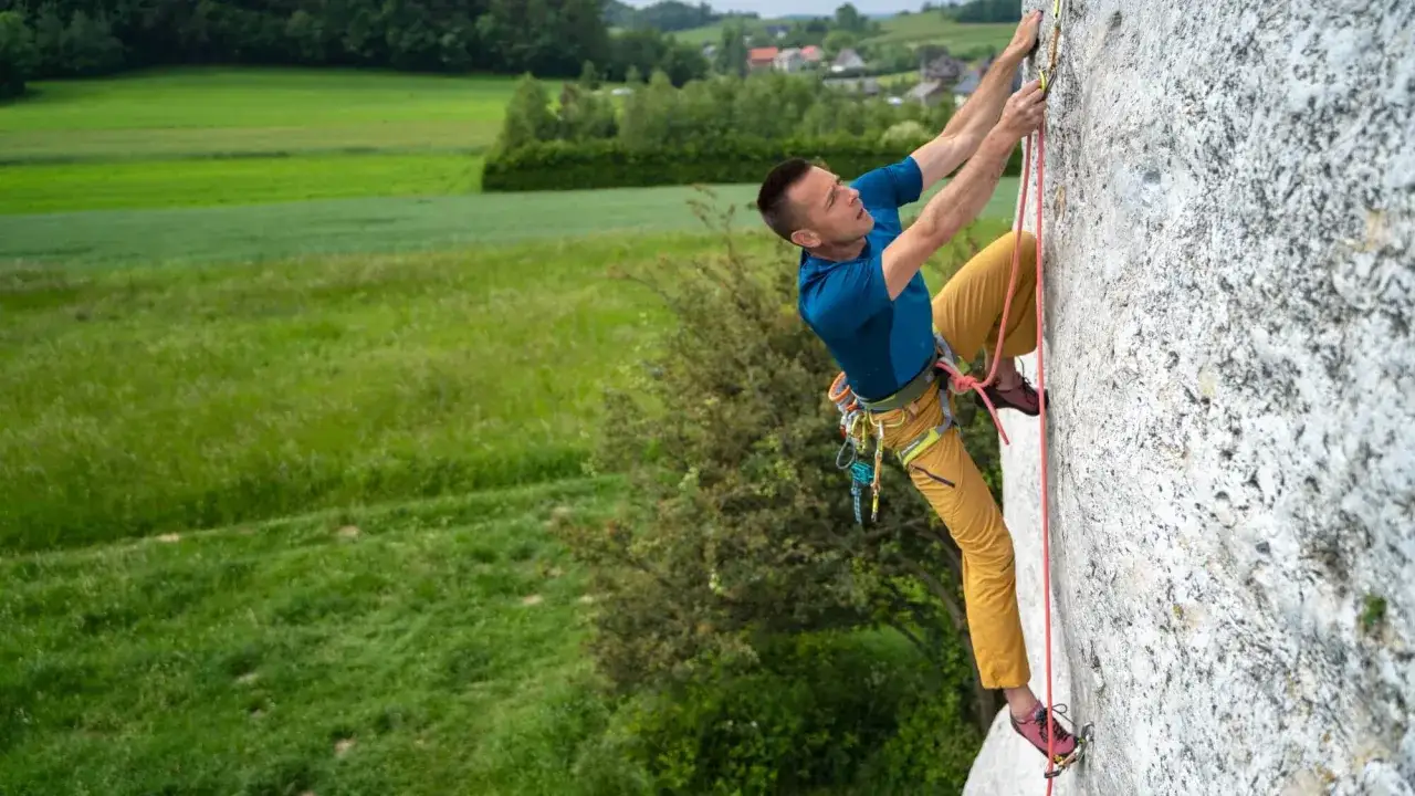 Mężczyzna wspina się po skale, używając liny i sprzętu. To klasyczna wspinaczka tradowa na tle zielonych p&oacute;l.