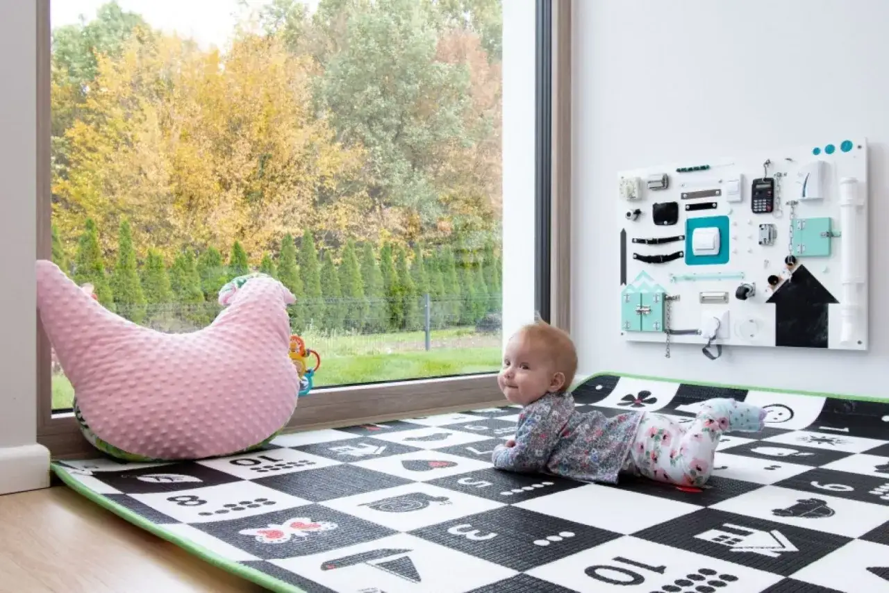 Urocze okno w pokoju dziecka aranżacje: maluch na macie obok r&oacute;żowej poduszki w kształcie kurczaka, za oknem jesienny ogr&oacute;d.