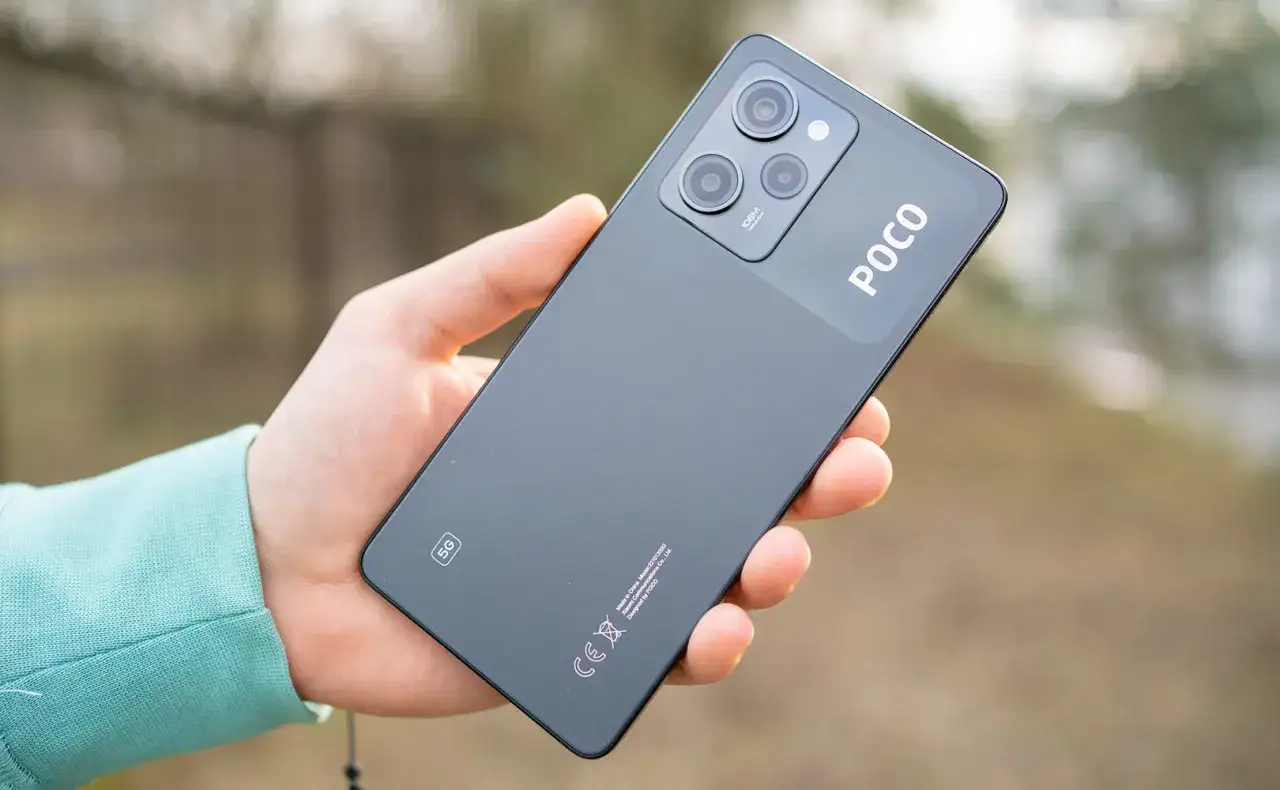 Dłoń trzyma ciemnoszary telefon POCO X5 5G z potr&oacute;jnym aparatem i logo POCO.
