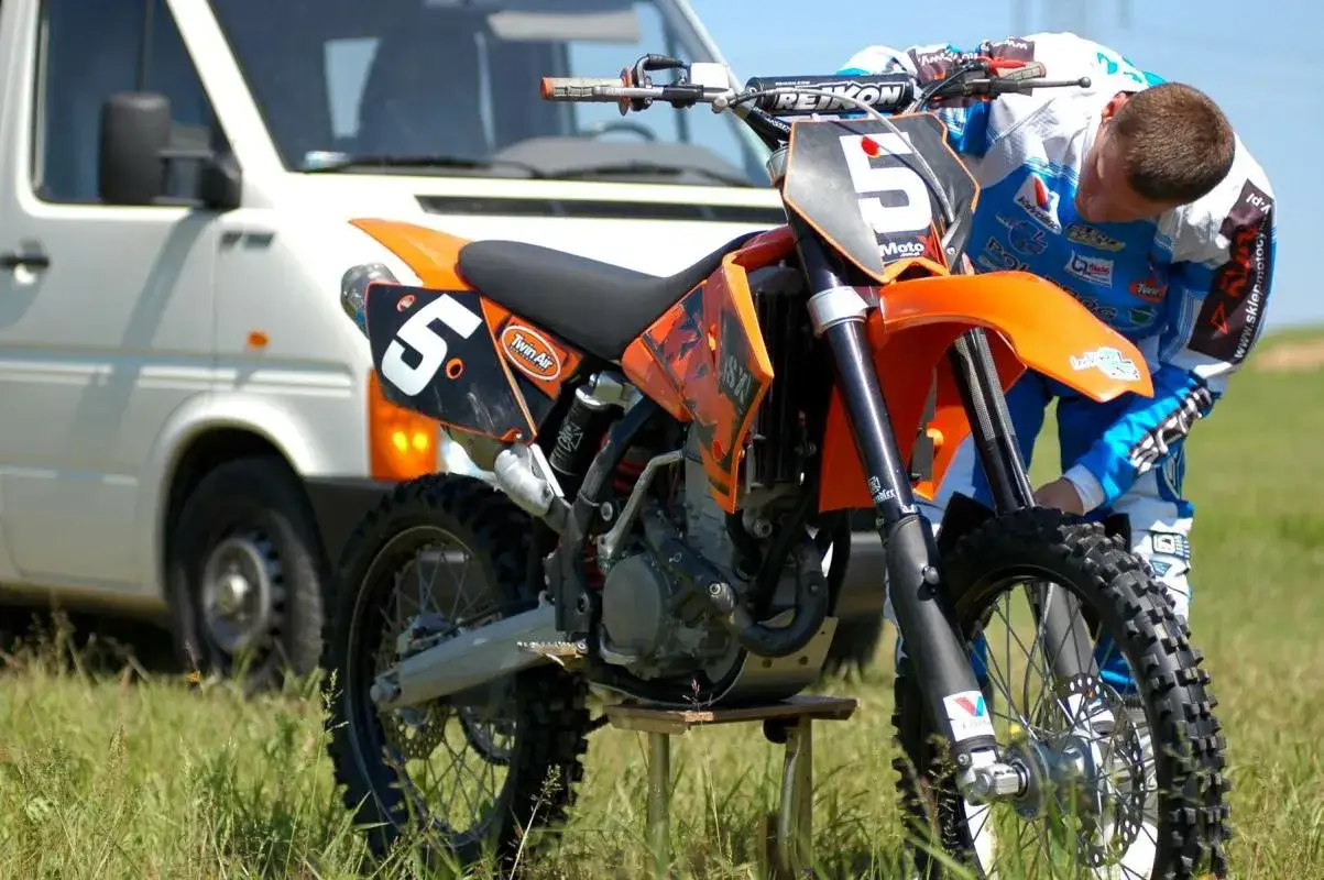 Pomarańczowy motocykl cross z numerem 5, gotowy do wyścigu. Kierowca w stroju przygotowuje się do jazdy.