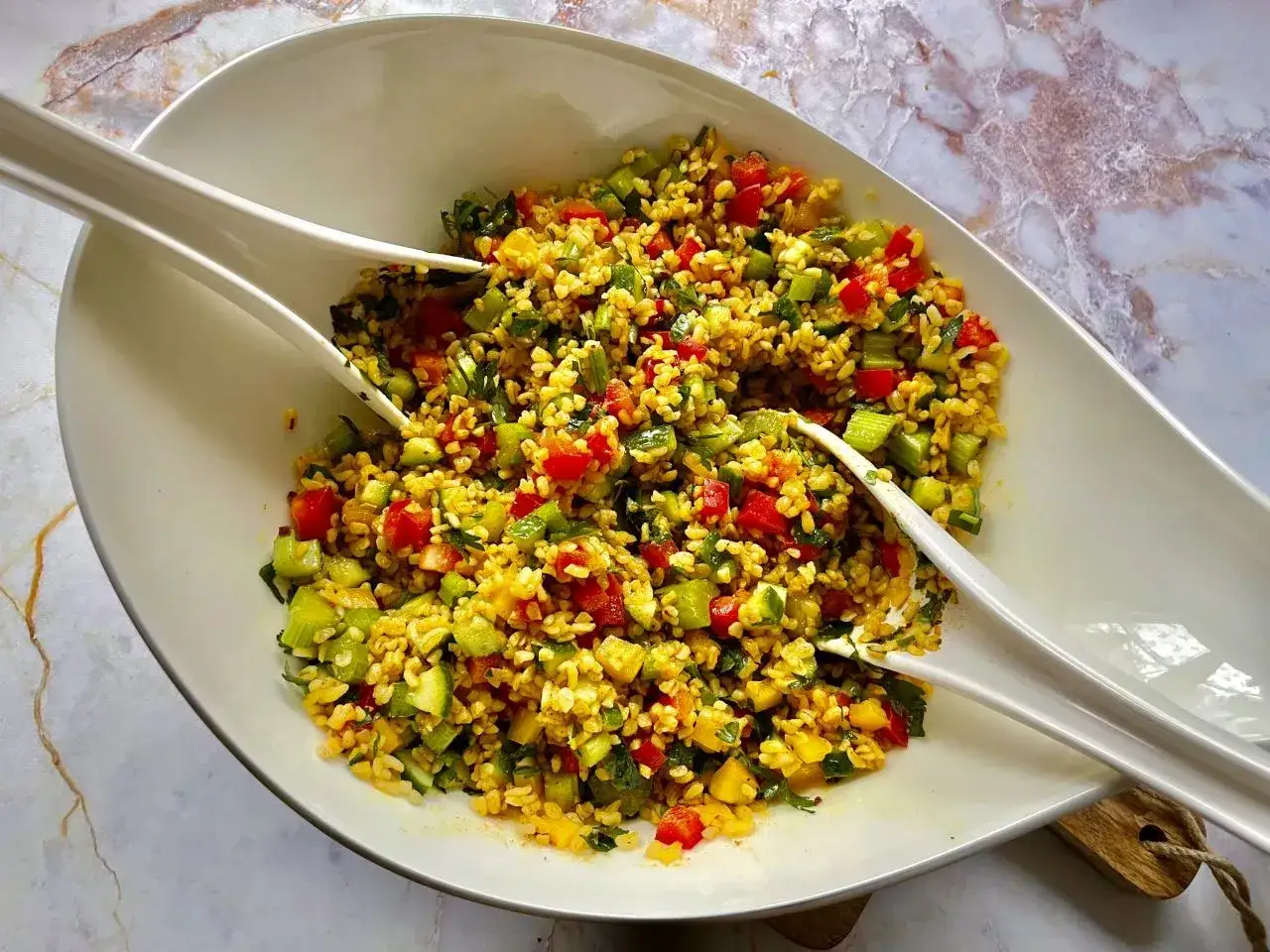 Kolorowa sałatka z kaszą bulgur, pomidorami, og&oacute;rkiem i ziołami, podana w białej misie z dwoma białymi szczypcami.