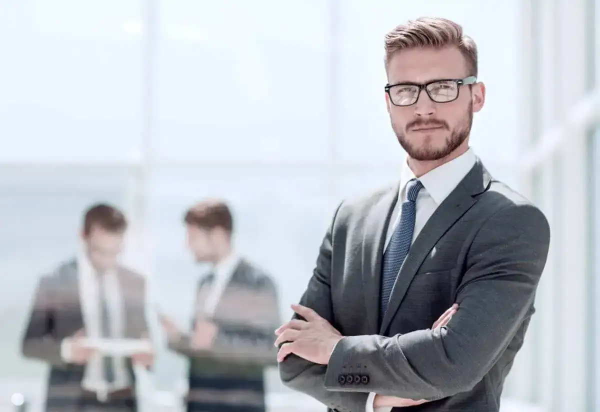 Młody mężczyzna w garniturze i okularach, stojący przed oknem. W tle dwaj inni mężczyźni. Czy to obraz tego, ile zarabia się w korporacji w Warszawie?