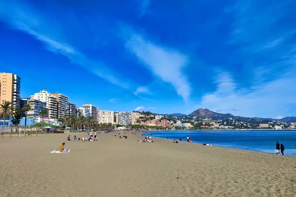 Piękna plaża w Maladze, gdzie widać, czy Malaga ma dostęp do morza. Ludzie odpoczywają na piasku, a w tle widać miasto i góry.