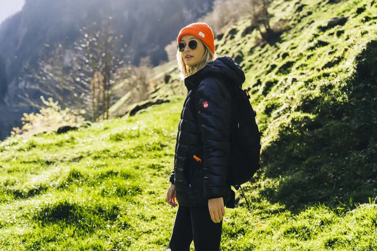 Kobieta w pomarańczowej czapce i czarnej kurtce z plecakiem na tle g&oacute;rskiego krajobrazu. Idealny zestaw, by wiedzieć, co spakować w g&oacute;ry.