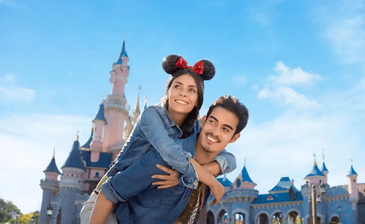 Szczęśliwa para z uszami Myszki Miki na tle zamku Disneya. Sprawdź, jaka jest bilet Disneyland cena i zaplanuj swoją magiczną podróż!