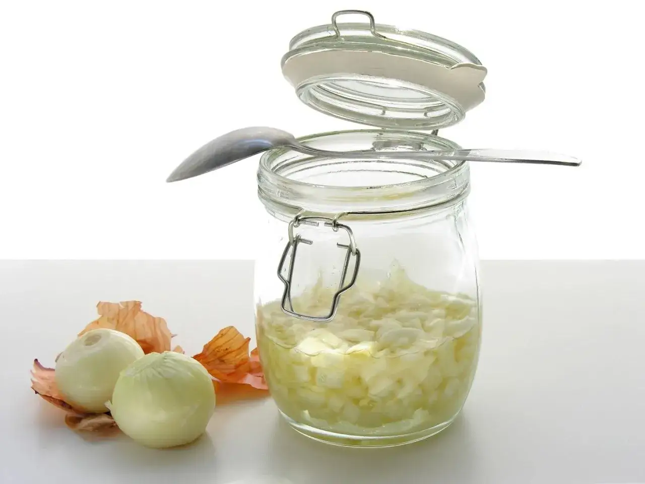 Słoik z domowym syropem z cebuli, obok dwie cebule i łuski. Łyżeczka opiera się o brzeg słoika.