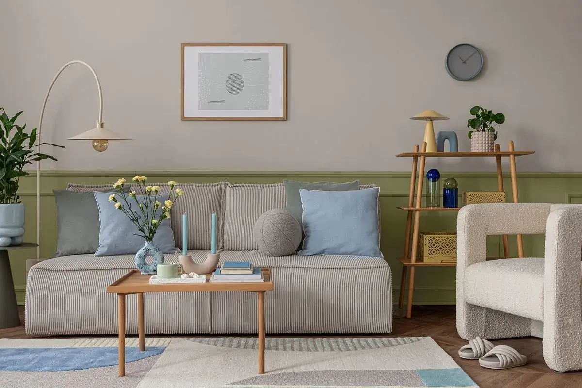 Przytulny salon z kanapą w paski, poduszkami w odcieniach błękitu i zieleni, stolikiem kawowym i regałem. Dominujący **kolor szałwiowy** na ścianie i zielona lamperia tworzą spokojną atmosferę.