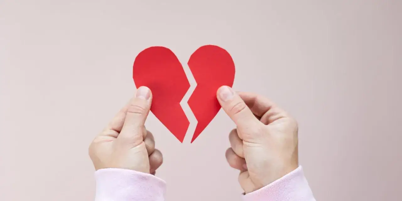 Dwie dłonie trzymają rozdartą na p&oacute;ł czerwoną kartkę w kształcie serca, symbolizującą złamane serce objawy.