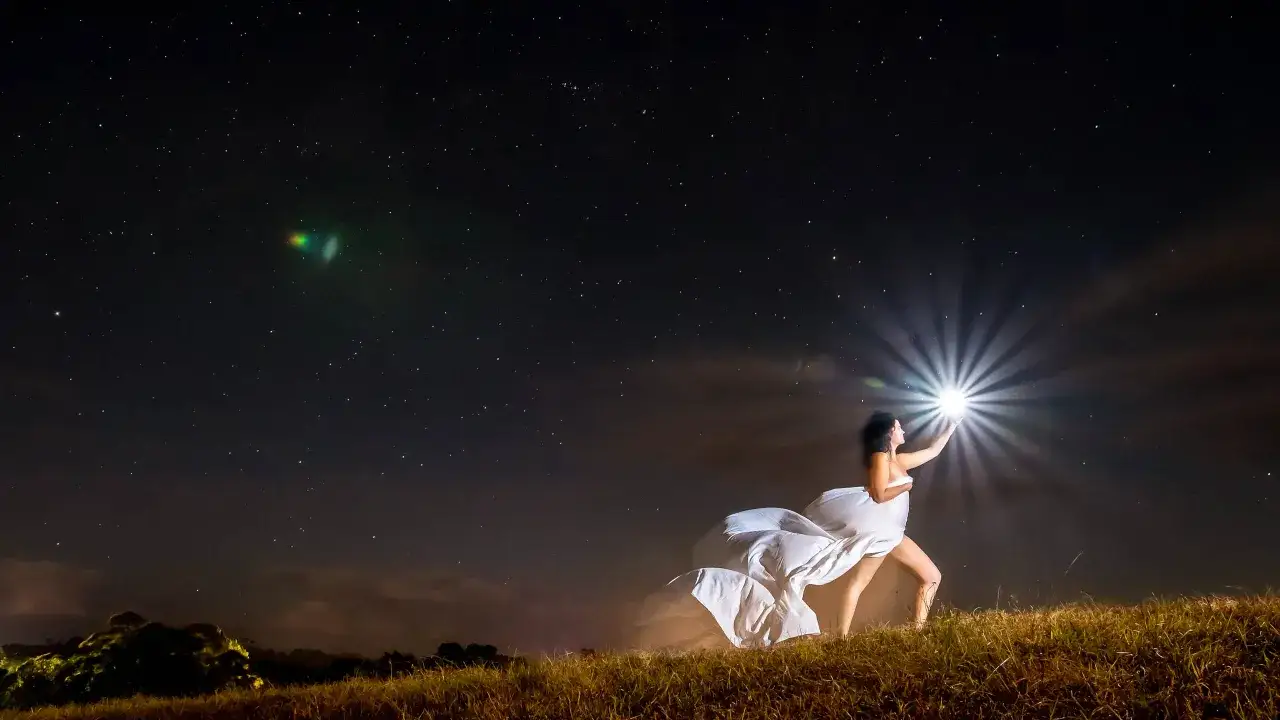Kobieta w bieli sięga po gwiazdę, kt&oacute;ra rozbłysła jak flash w aparacie na tle nocnego nieba pełnego gwiazd.