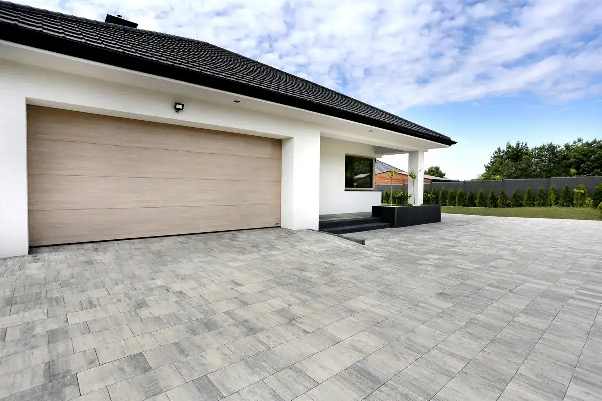 Nowoczesny podjazd do domu z betonowej kostki, prowadzący do garażu z jasną bramą.