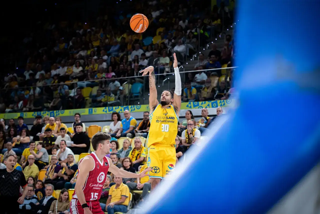 Jugador del club baloncesto Gran Canaria lanza a canasta ante un rival. El público anima en las gradas.