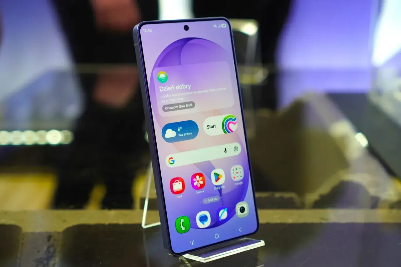 Smartfon z interfejsem One UI 8.5, prezentujący ekran gł&oacute;wny z widżetem pogody i ikonami aplikacji.