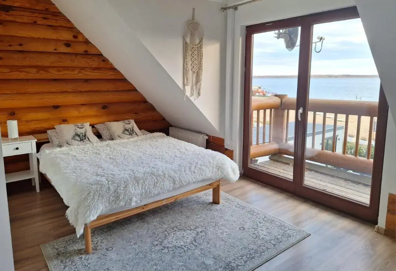 Przytulny domek nad jeziorem Śląsk. Sypialnia z widokiem na wodę, drewniane ściany i miękkie futrzane koce.