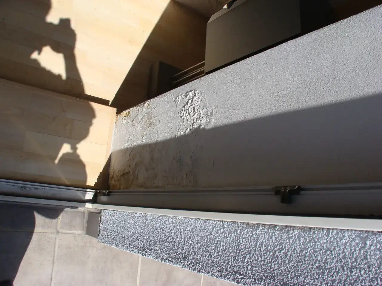 Balkon w bloku z wielkiej płyty, z widocznymi śladami wilgoci i łuszczącej się farby na ścianie. W tle drewniana podłoga i cień osoby trzymającej aparat.