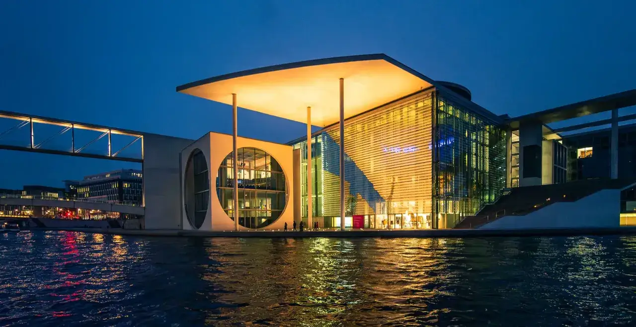 Nowoczesny budynek nad wodą, z podświetlanym dachem i szklanymi fasadami, to muzeum sztuki nowoczesnej w Berlinie.