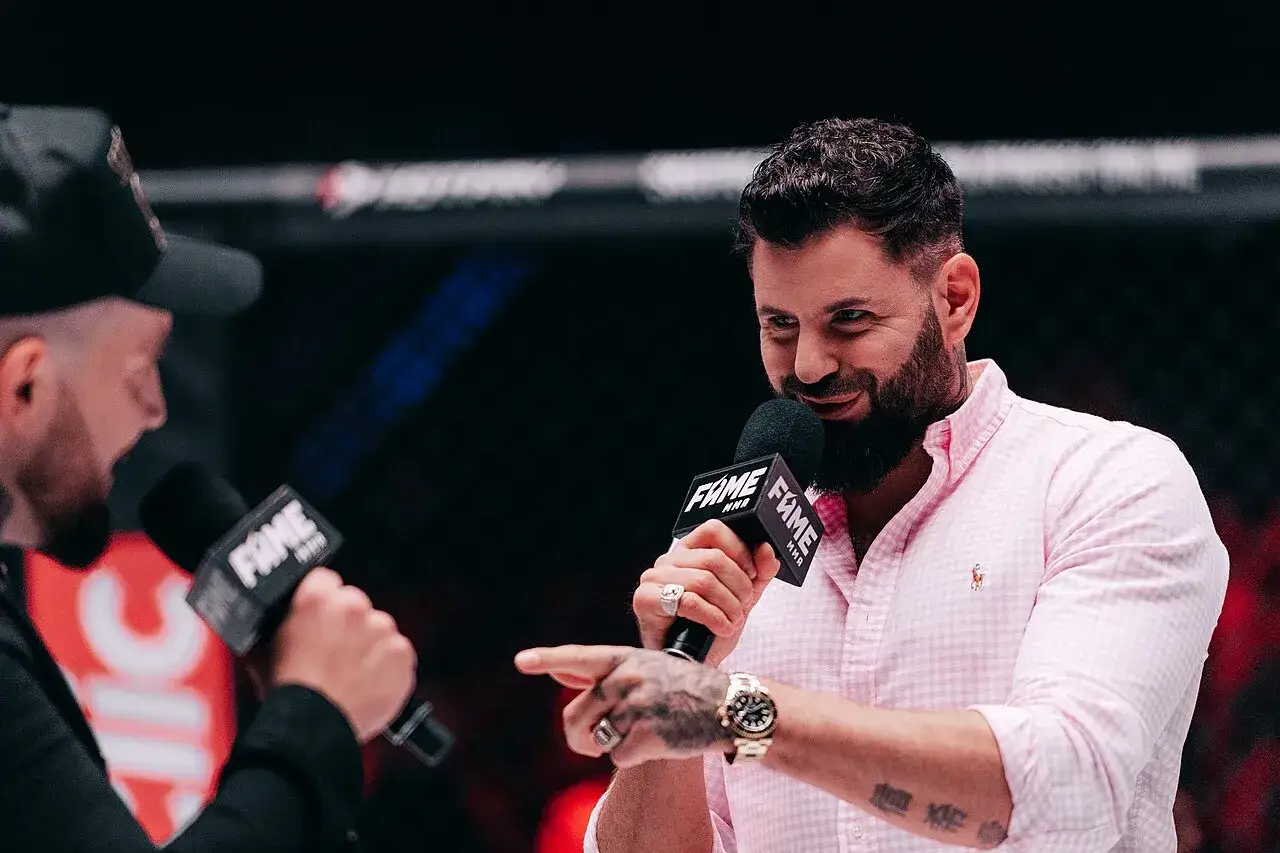 Wojtek Gola, znany z FAME MMA, w różowej koszuli, z mikrofonem i tatuażami.