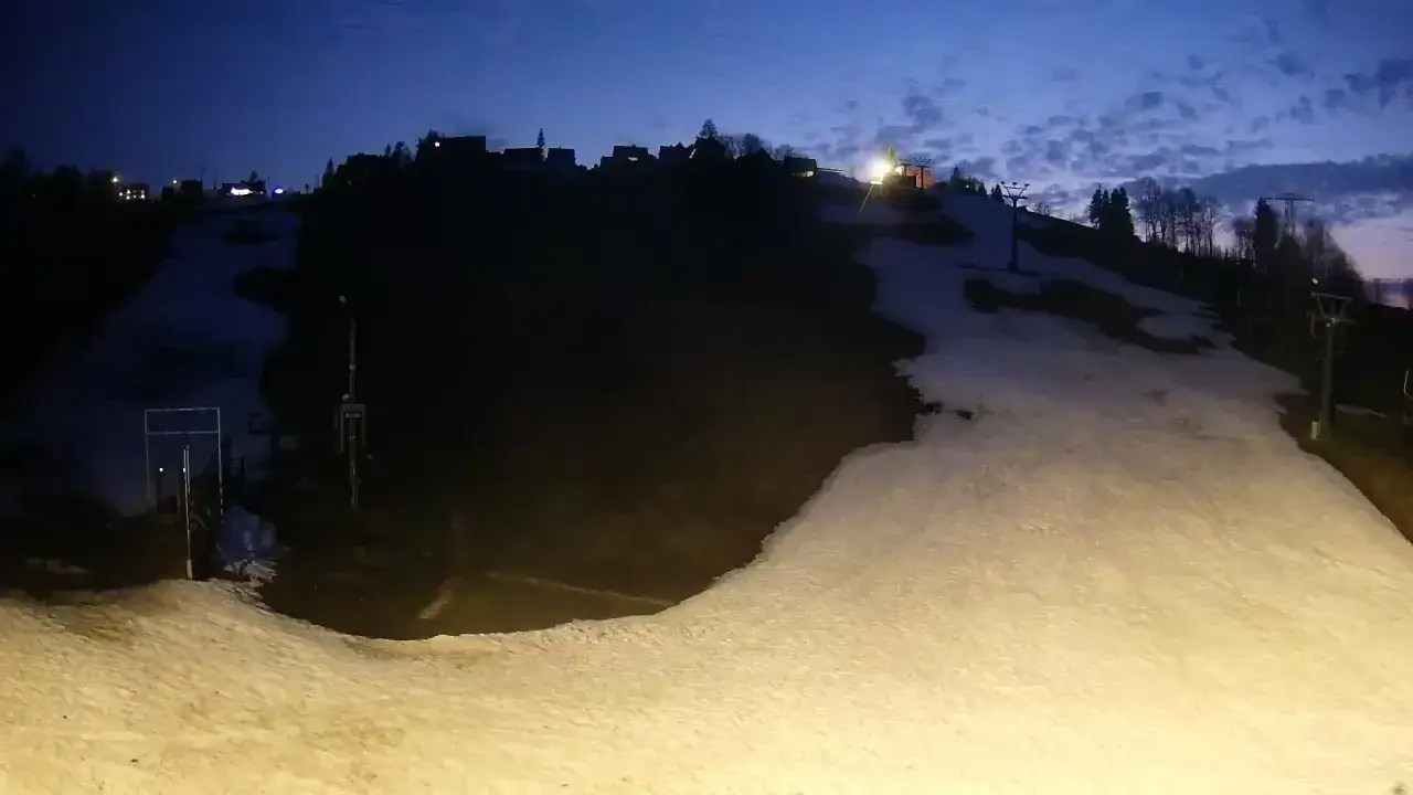 Bukowina Tatrzańska stok kamera nocą. Widać ośnieżony stok, wyciąg narciarski i zabudowania na wzg&oacute;rzu.