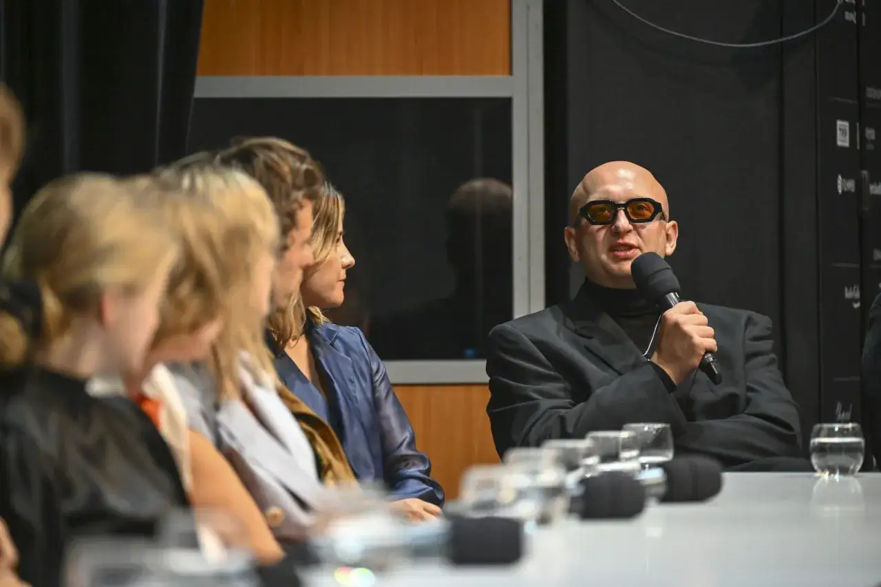 Nikodem Chabior w okularach i czarnym golfie przemawia do mikrofonu, siedząc przy stole z innymi osobami.