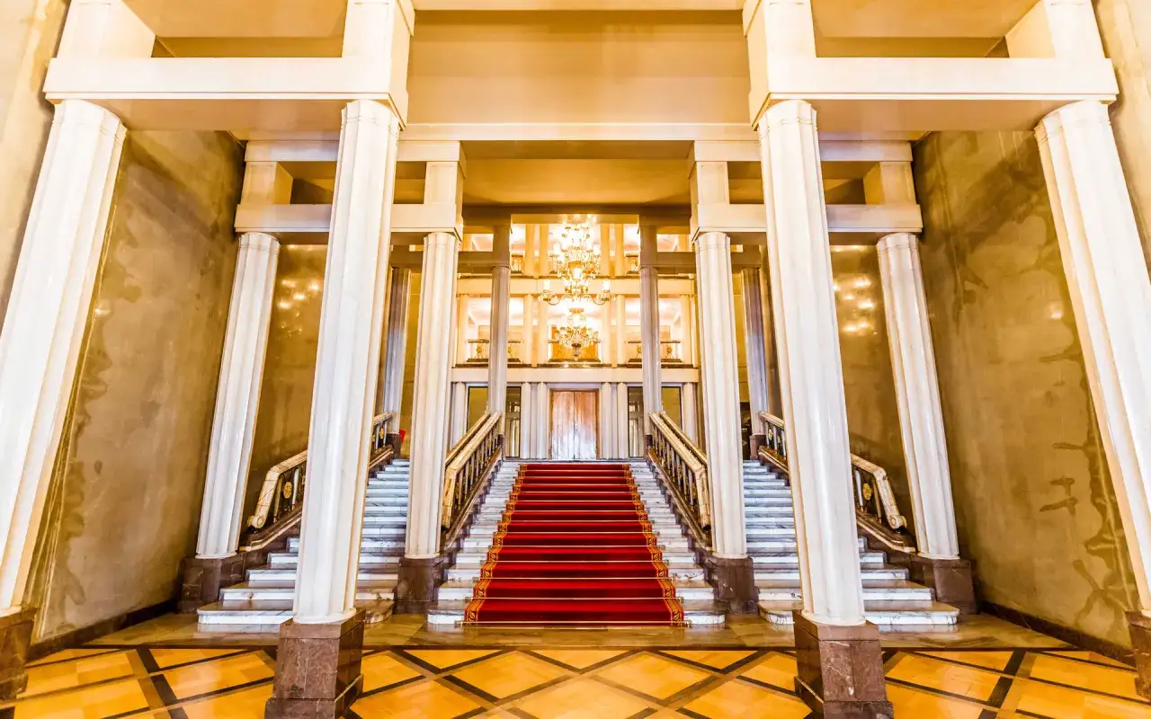 Eleganckie foyer teatru z czerwoną wykładziną na schodach, marmurowymi kolumnami i kryształowym żyrandolem.