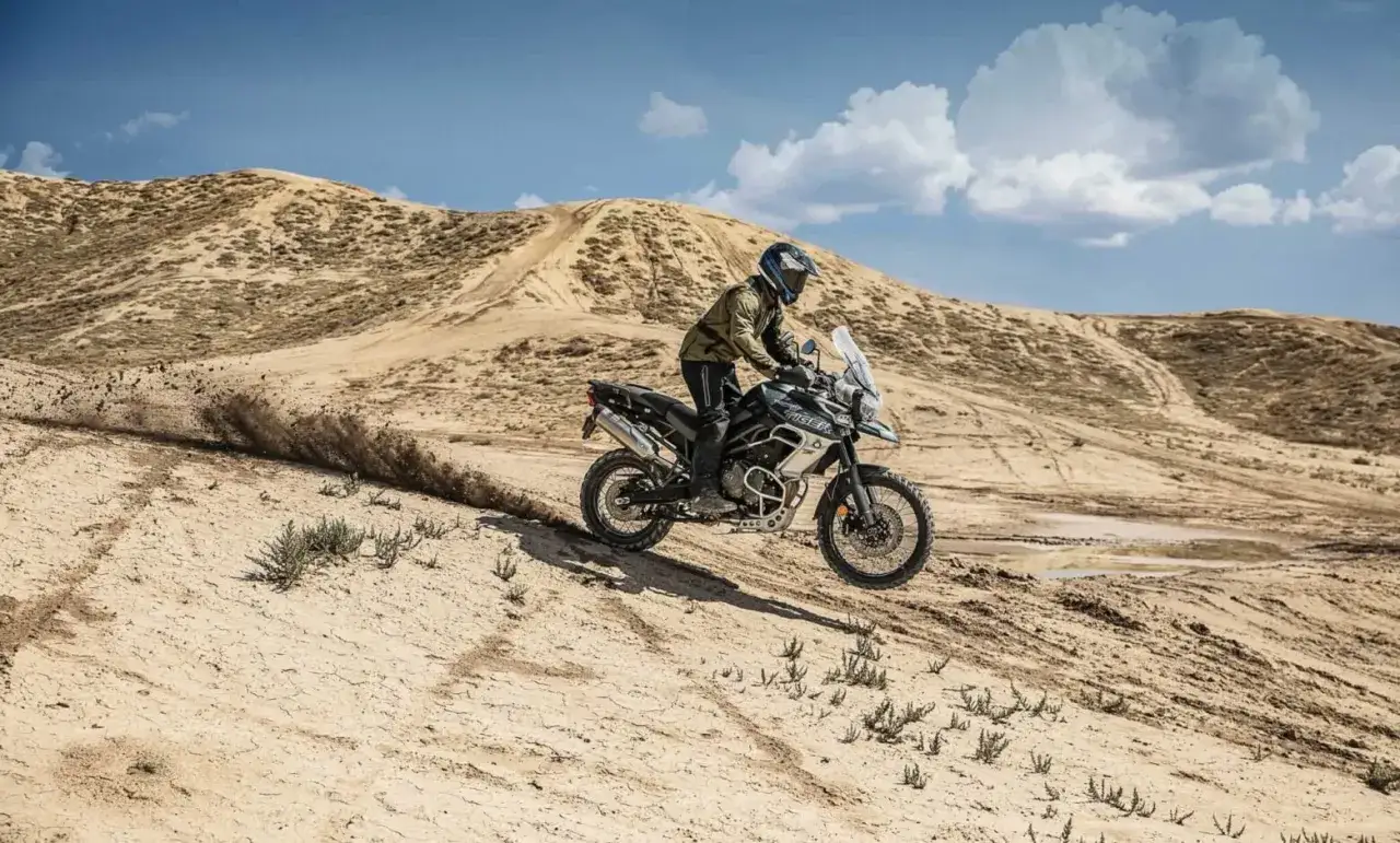 Motocyklista na Triumph Tiger 800 XC wjeżdża na piaszczyste wzg&oacute;rze, unosząc tumany kurzu. Dane techniczne tego modelu pozwalają na takie wyzwania.