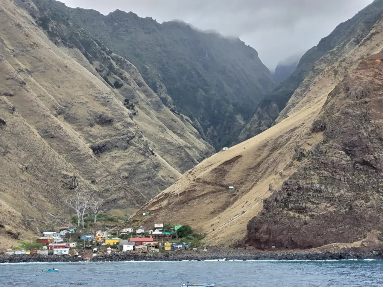 Osiedle w dolinie, przypominające przypadki Robinsona Crusoe, z kolorowymi domkami i łodziami na morzu u podnóża stromych, trawiastych gór.