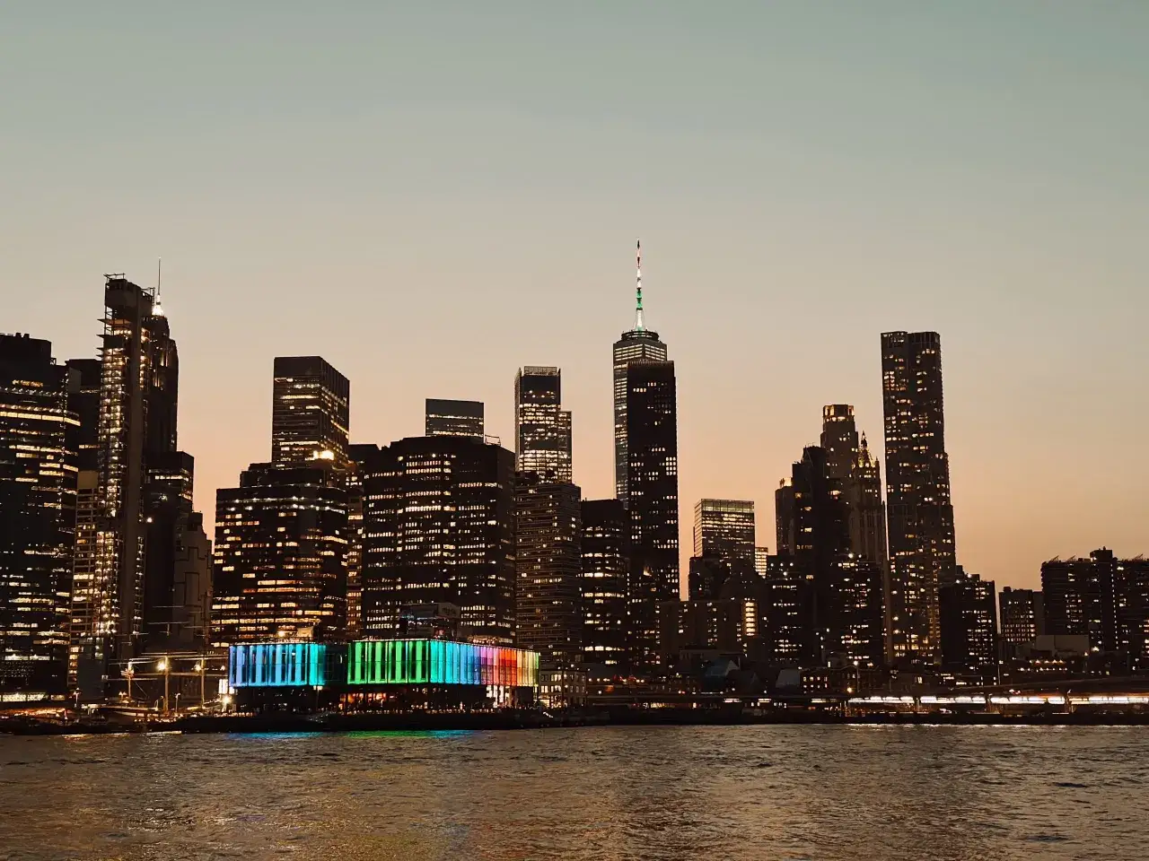 Widok na nocny Nowy Jork z rzeki. Widać oświetlone wieżowce, w tym One World Trade Center. Kolorowy budynek na pierwszym planie dodaje uroku tej wycieczce.