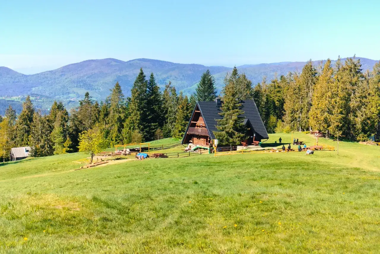 Malownicza bac&oacute;wka Krawc&oacute;w Wierch otoczona lasem i zielonymi łąkami, z widokiem na g&oacute;ry.