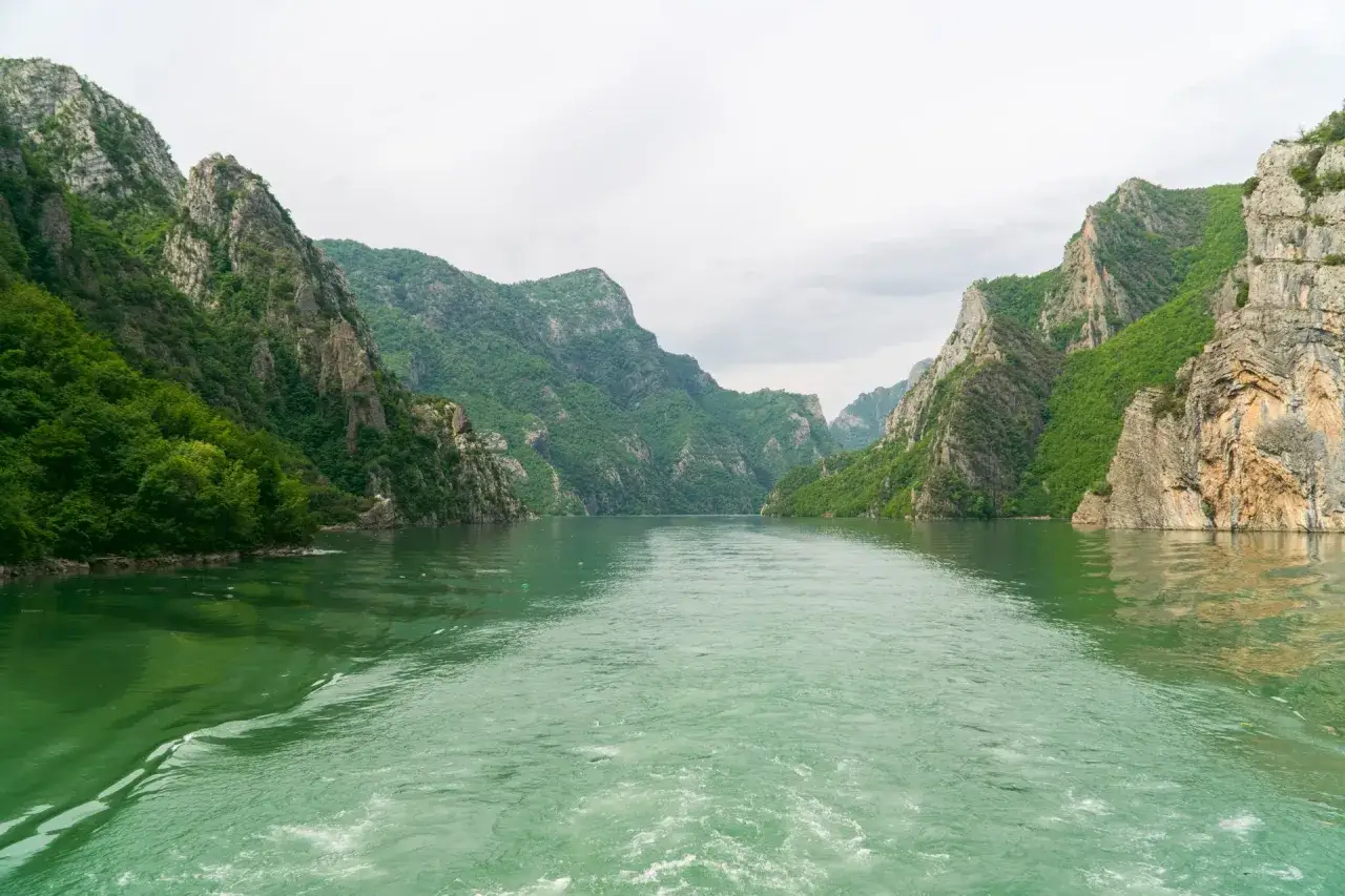 Widok na zieloną rzekę w otoczeniu stromych, skalistych g&oacute;r porośniętych bujną zielenią. To malownicza trasa, kt&oacute;ra może być częścią podr&oacute;ży, jak dojechać do Albanii.